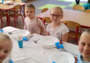 Dzieci siedzące przy stole nakrytym białym obrusem. Na stole ustawione talerze ze sztućcami i dekoracja z serwetek niebieskich.